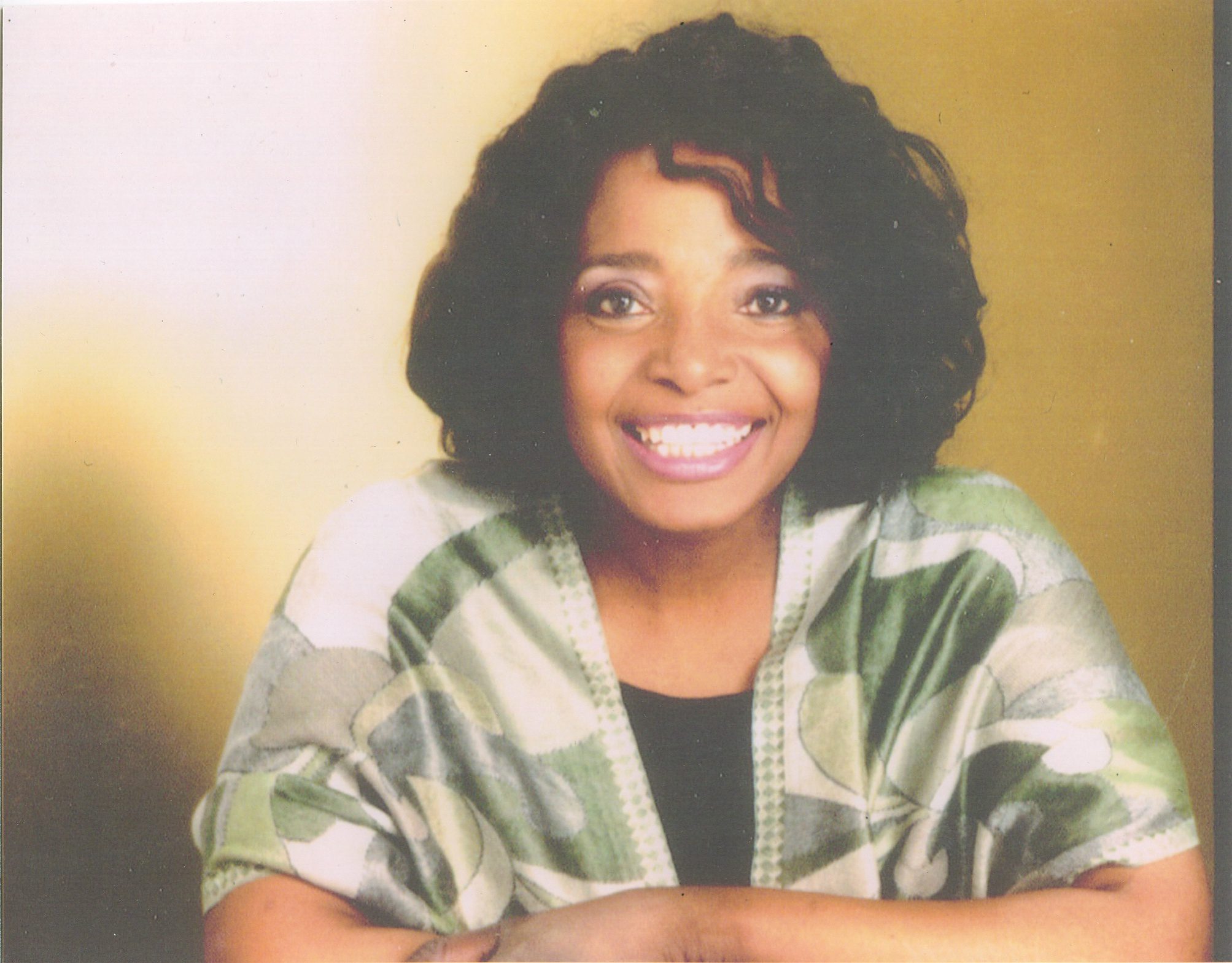 Smiling woman with curly hair wearing a green and white patterned top.